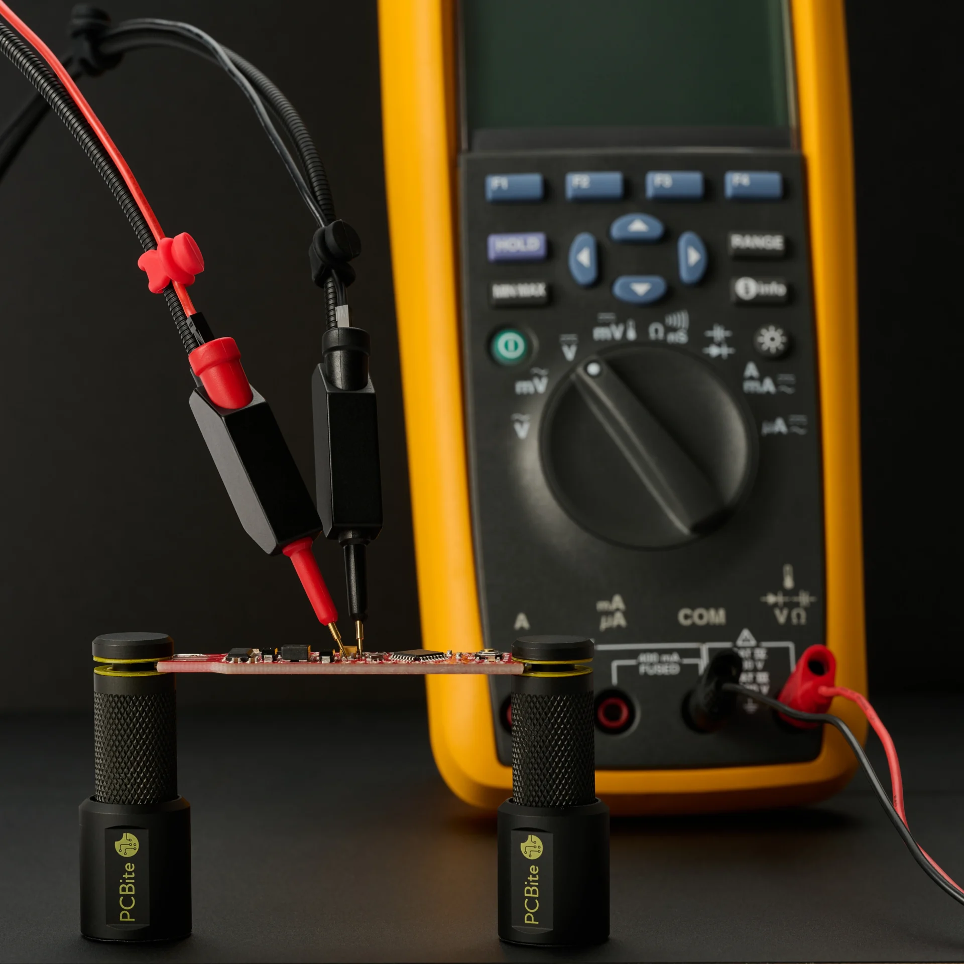 PCB in PCBite holders, wired to a FLUKE multimeter so the readings need no hands.