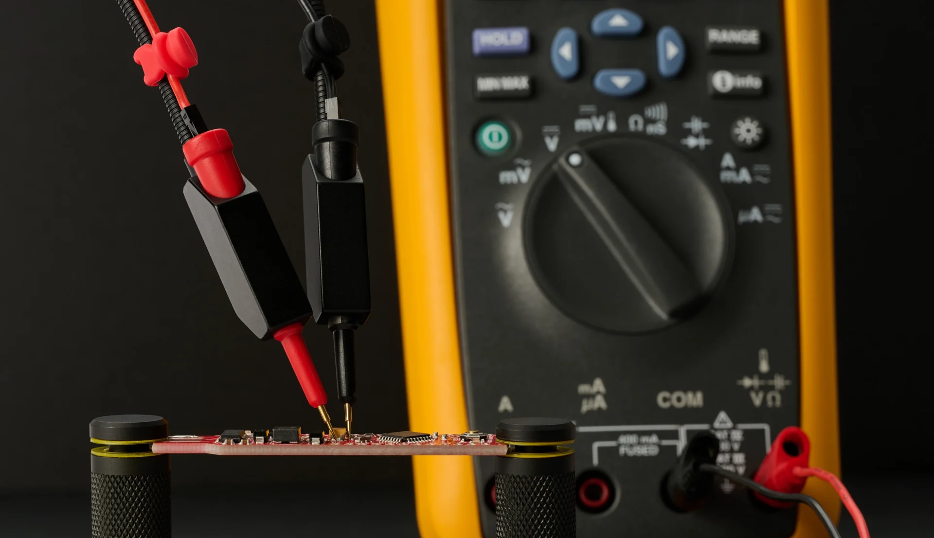 Red and black handsfree probes measuring a small PCB in PCBite holders, wired to a multimeter in the background.
