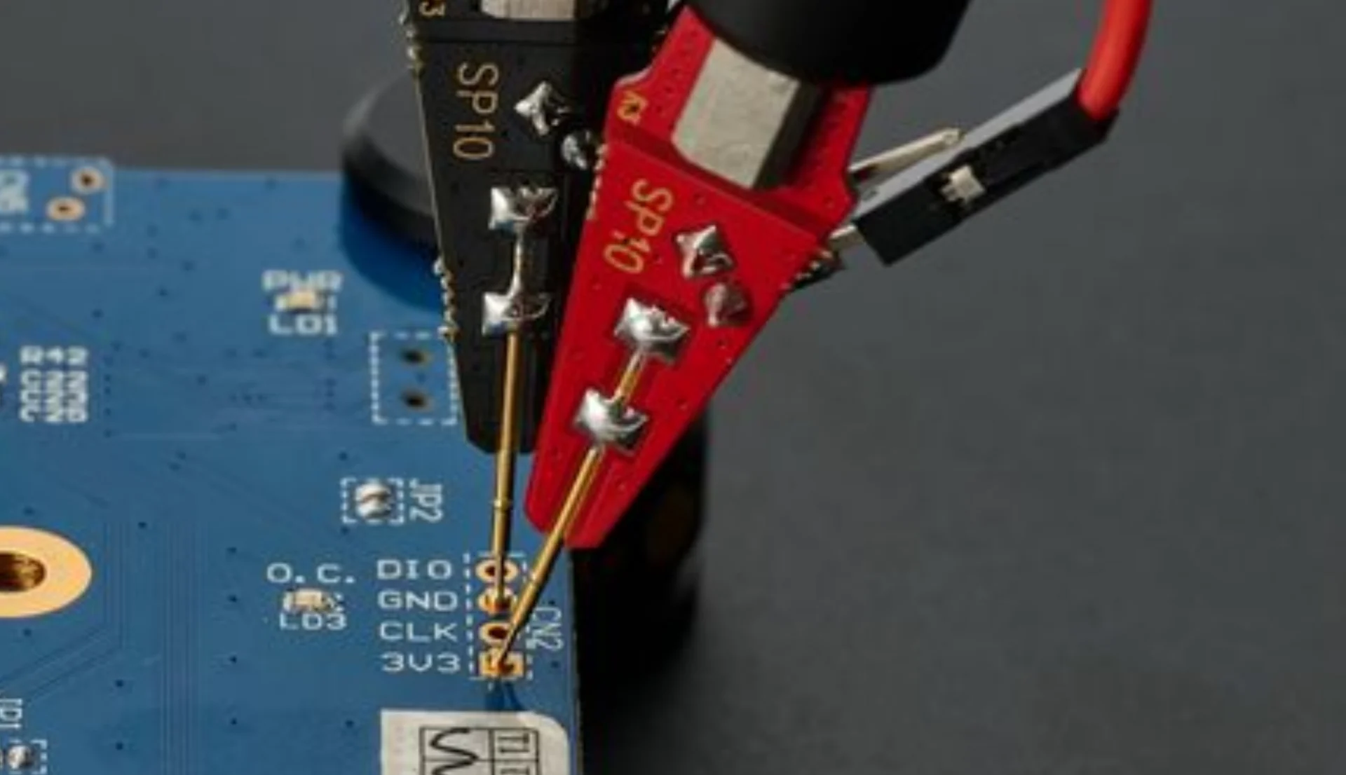 Red and black SP10 probes with gold spring-loaded needles resting on the pins of a small header on a blue PCB.