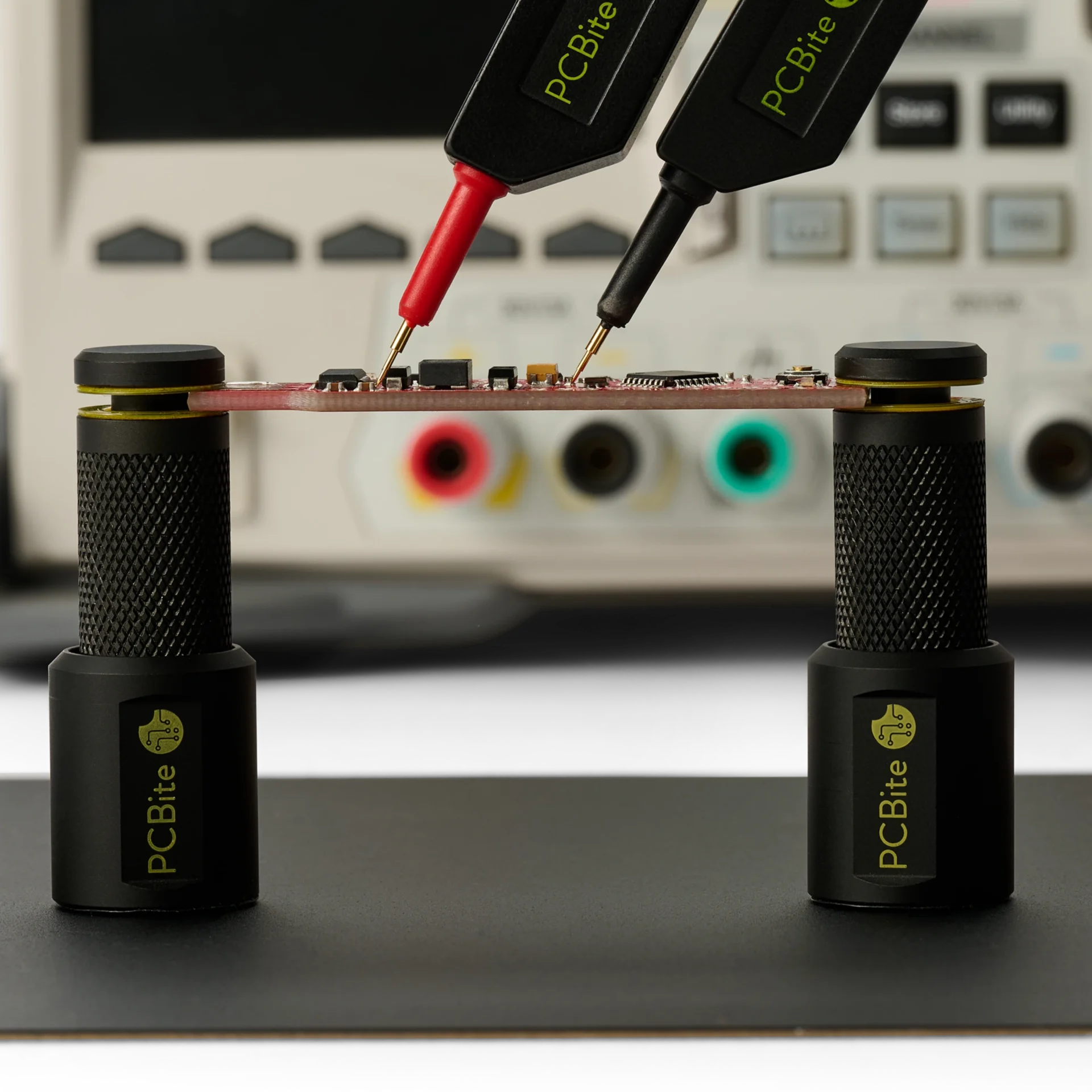 Powered PCB held by two PCBite holders with yellow insulation washers, probed with red and black probes in front of a bench supply.