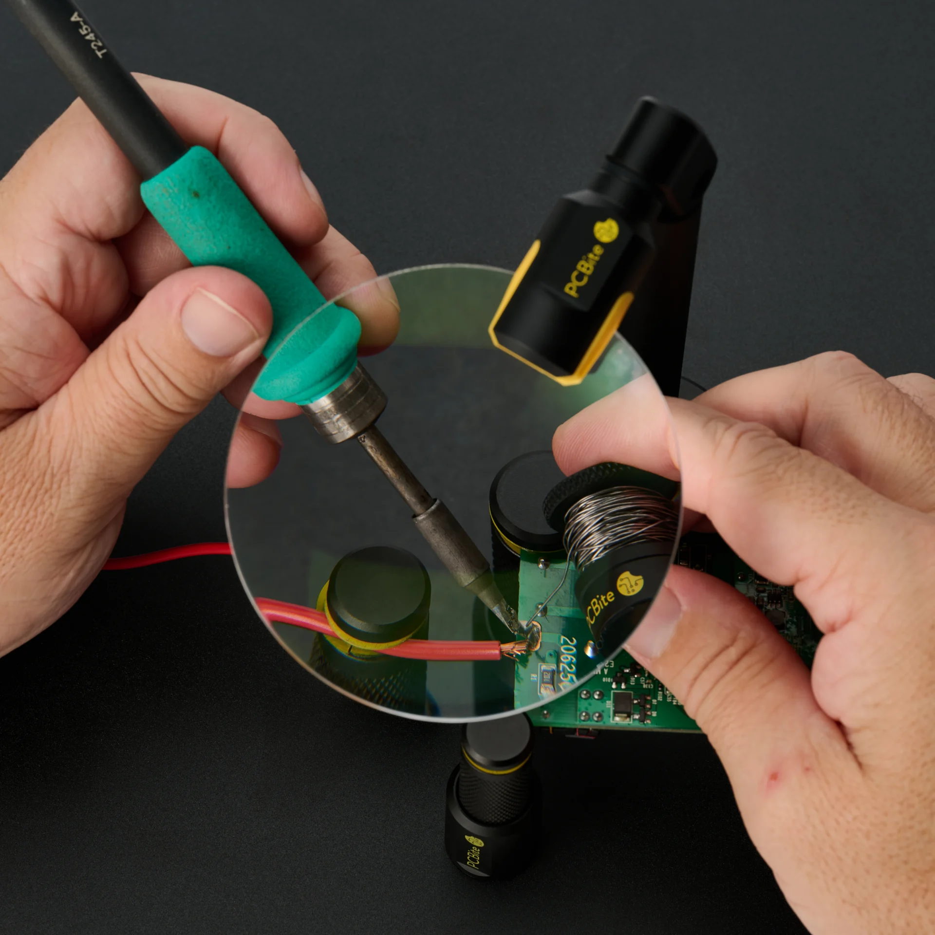 Soldering a red wire to a PCB through the PCBite magnifier, with PCBite holders keeping the wire and the board steady.