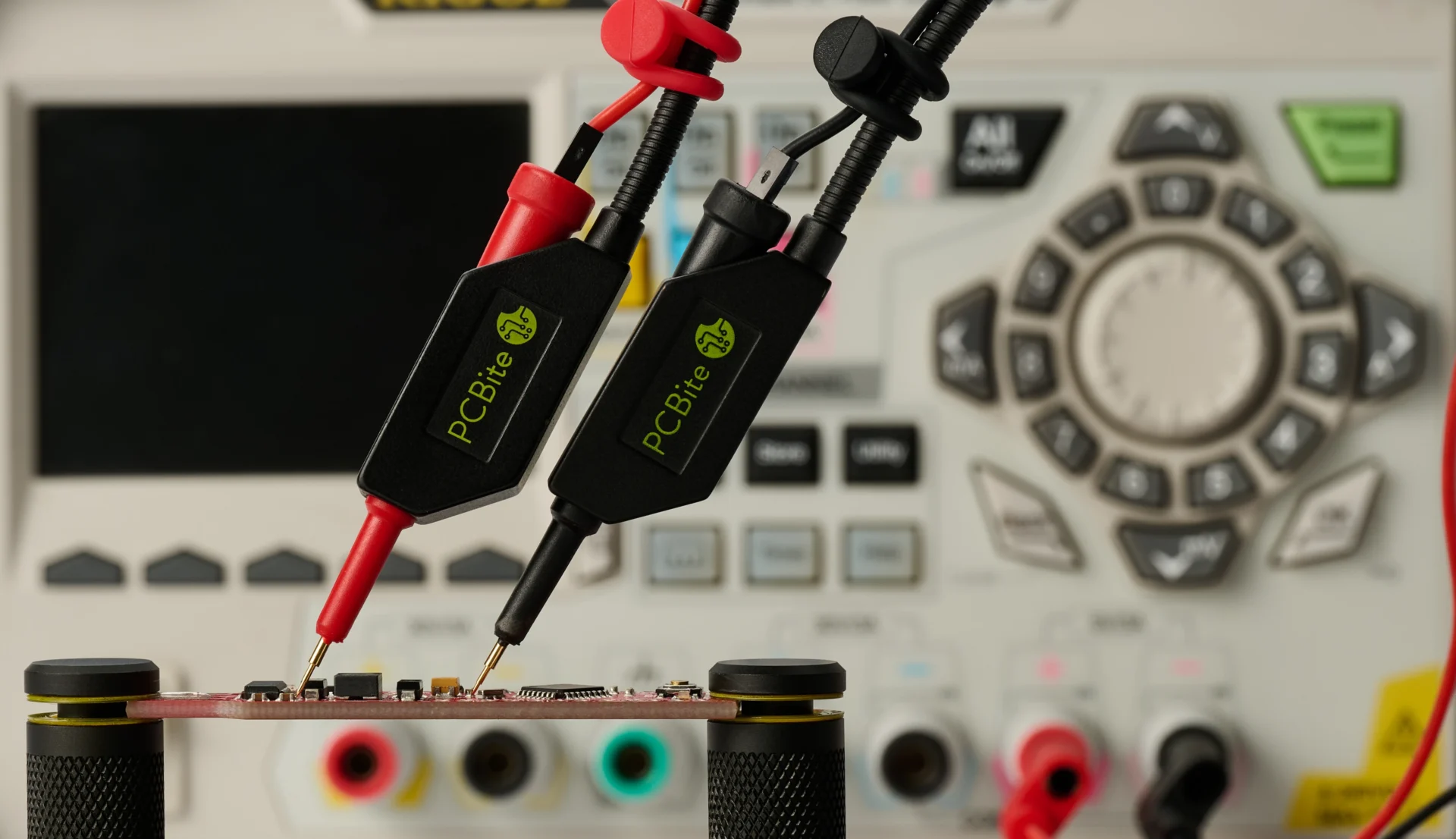 Red and black handsfree probes on a PCB between two PCBite holders, connected to the bench power supply behind.